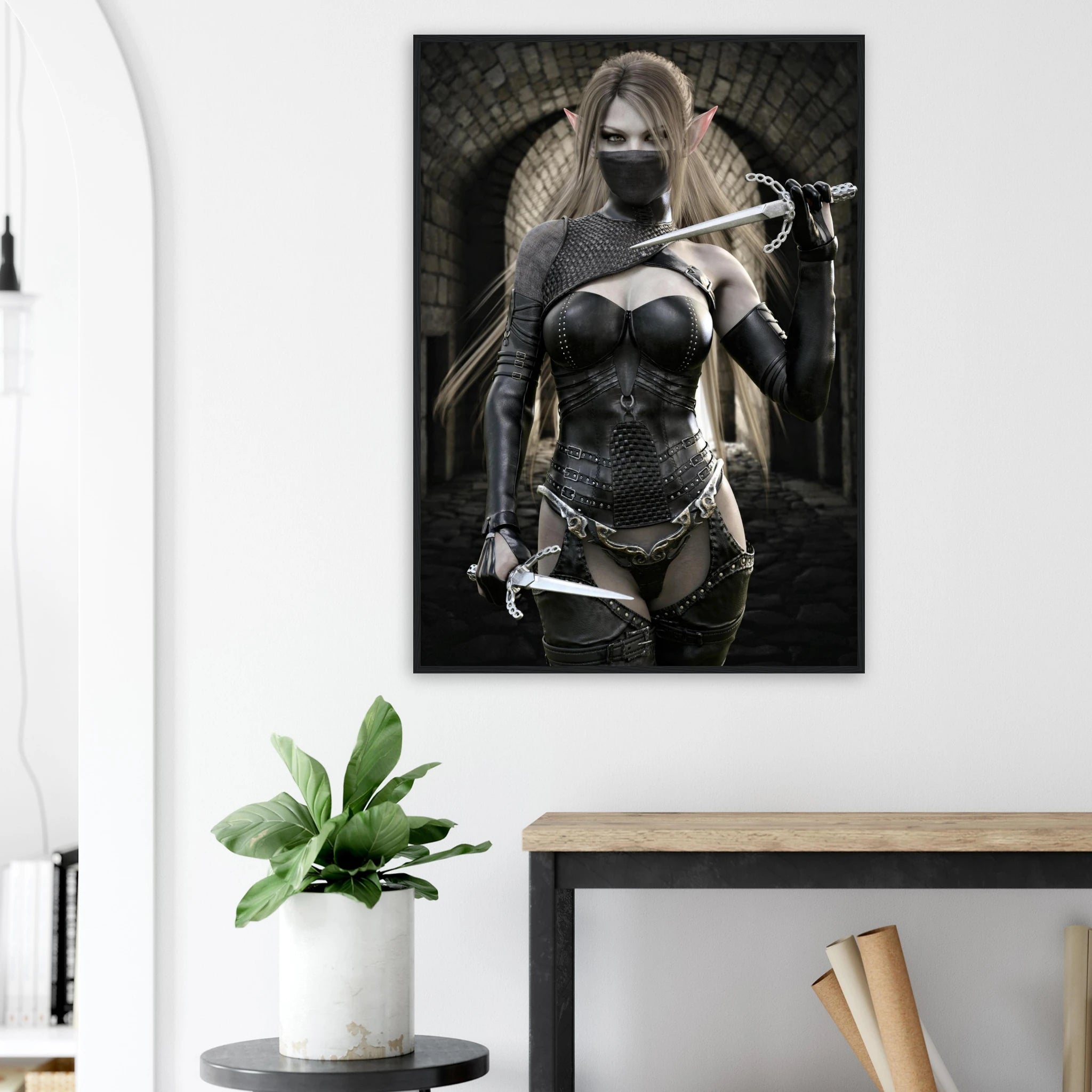 A framed vertical wall art print showing a fantasy female warrior in dark chainmail-style armor holding swords, displayed on a white wall above a console table with a potted plant.