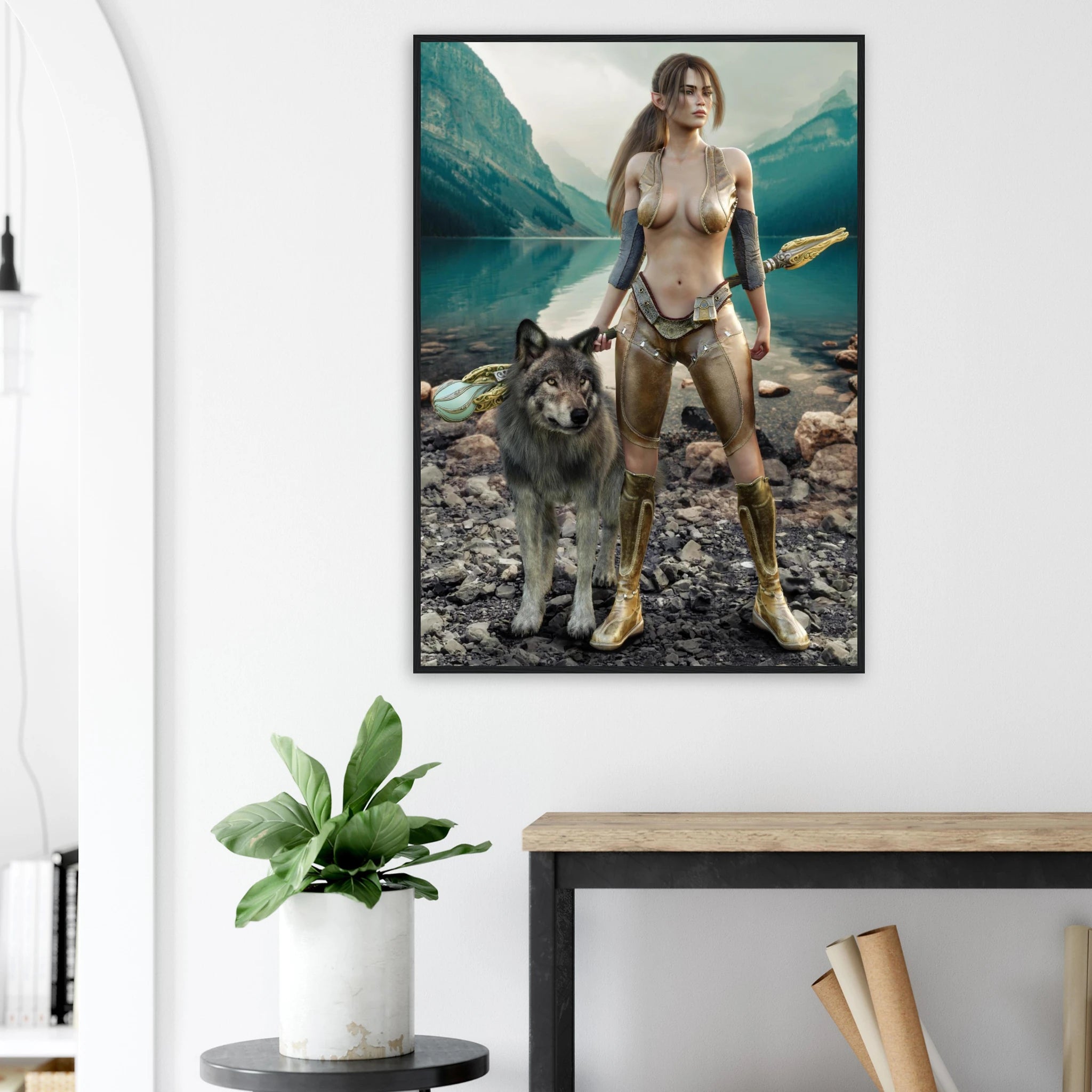 A framed vertical art print hanging on a white wall above a wooden table, showing a fantasy-style scene of a nude woman with a wolf in front of a mountain lake. The room includes a potted plant and part of a chair.